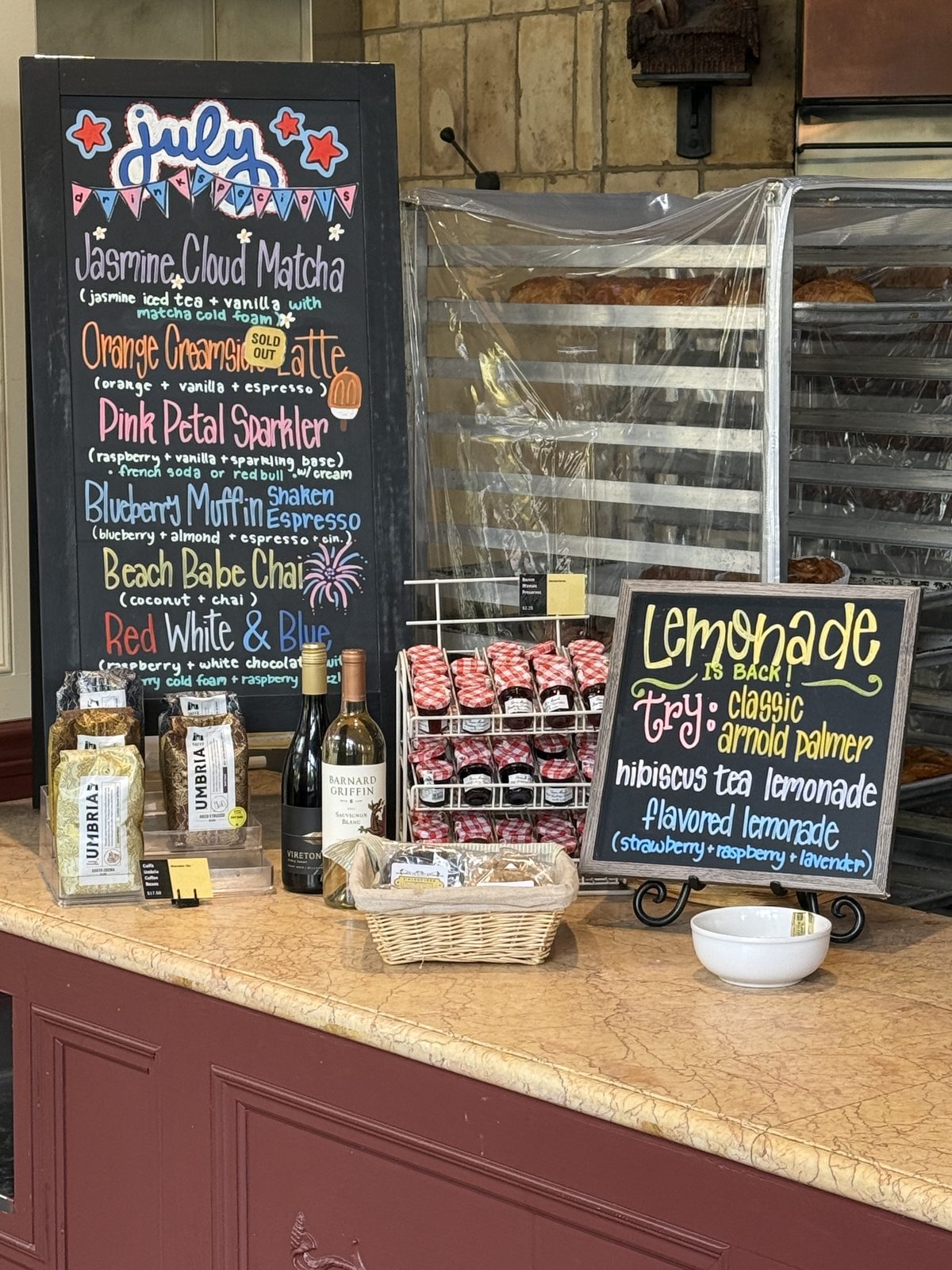 A hand-chalked specials board reading July, listing seasonal drinks like Jasmine Cloud Matcha, Pink Petal Sparkler and Beach Babe Chai, next to jars of jam and bottles of wine for sale on the counter