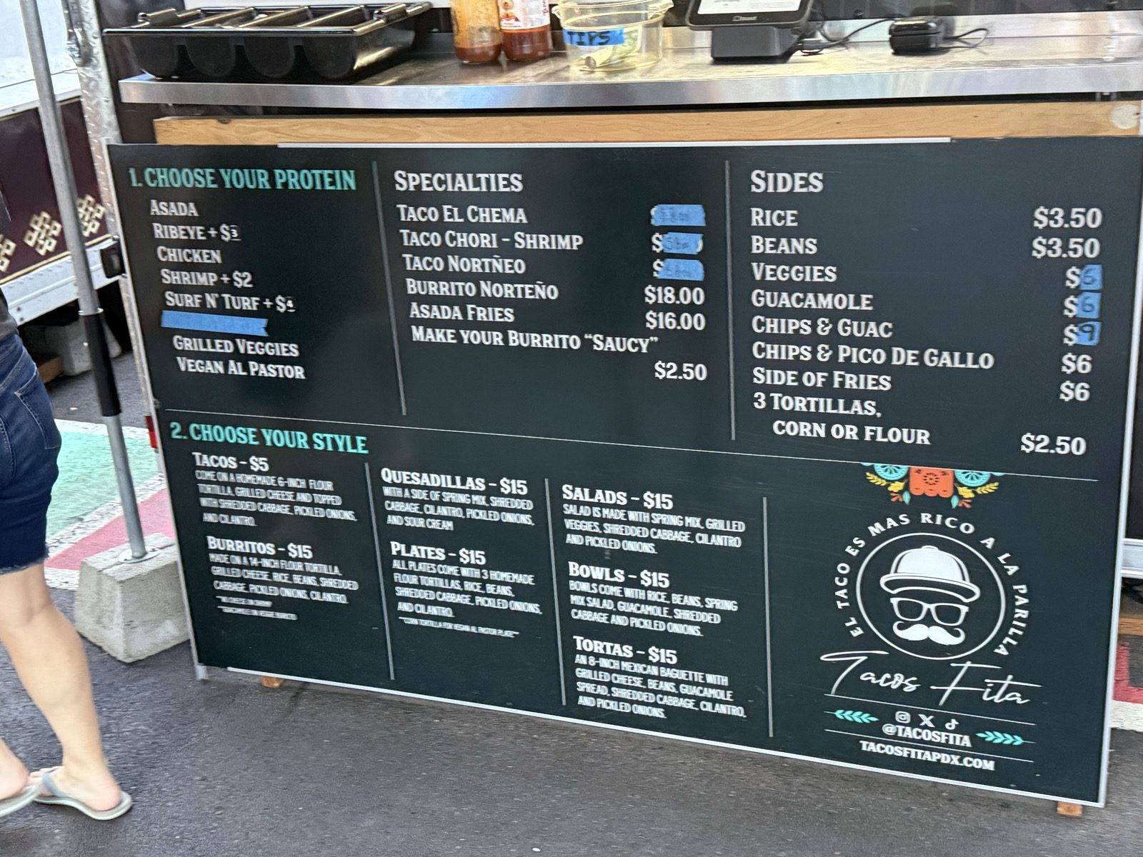The menu board at Tacos Fita, listing proteins including asada, ribeye, chicken and shrimp, tacos for five dollars on a homemade flour tortilla with grilled cheese, shredded cabbage and pickled onions, plus burritos, quesadillas and specialties