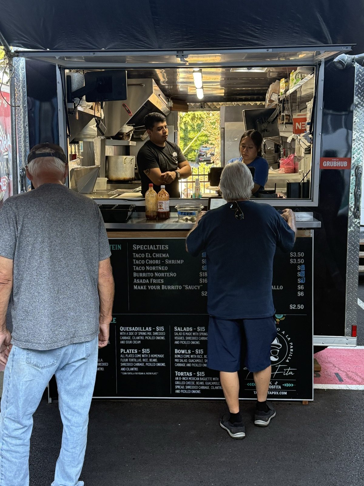 Two customers ordering at the walk-up window of the Tacos Fita food truck, with cooks working behind the counter and the menu board visible in the foreground