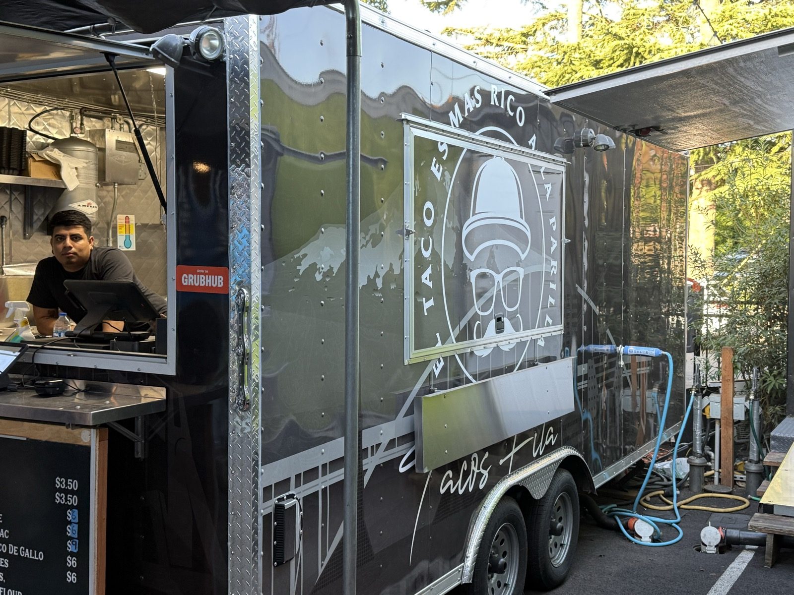 The Tacos Fita food truck in the Woodstock neighborhood of Portland, Oregon, a black trailer with a mustached man in a hat logo and the slogan El Taco Es Mas Rico A La Parilla, with a cook working behind the order window