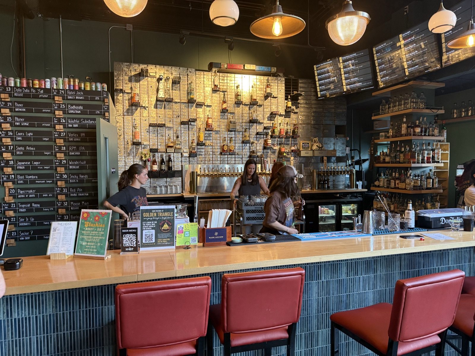 The full bar at The Heist in Portland, Oregon, with a long chalkboard beer menu on the left, bottles of liquor on shelves against a green tiled wall, and bartenders working behind a wooden counter with red leather stools