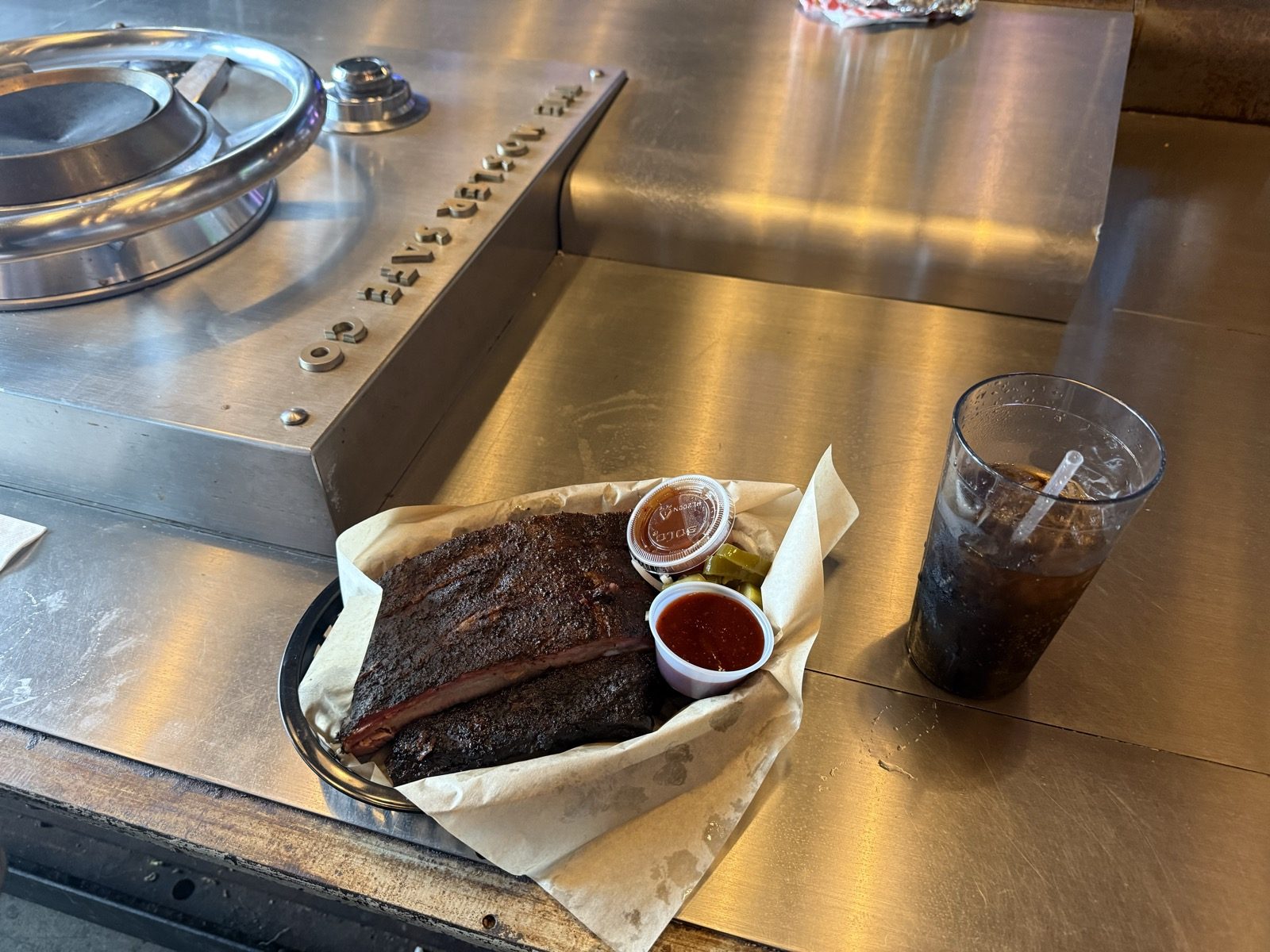 A rack of Bark City BBQ pork ribs and cups of pickles and barbecue sauce in a paper-lined basket, next to a glass of Coca-Cola, sitting on a stainless steel table built around the mechanism of an old bank vault door at The Heist in Portland, Oregon