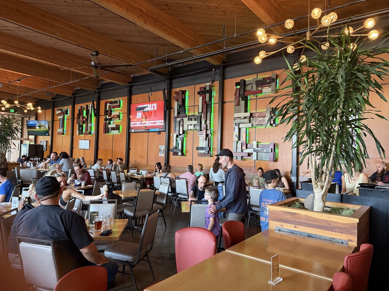 The indoor dining hall at The Heist in Portland, Oregon, with a high wood-beamed ceiling, long communal tables, and a large wall sculpture spelling out HEIST