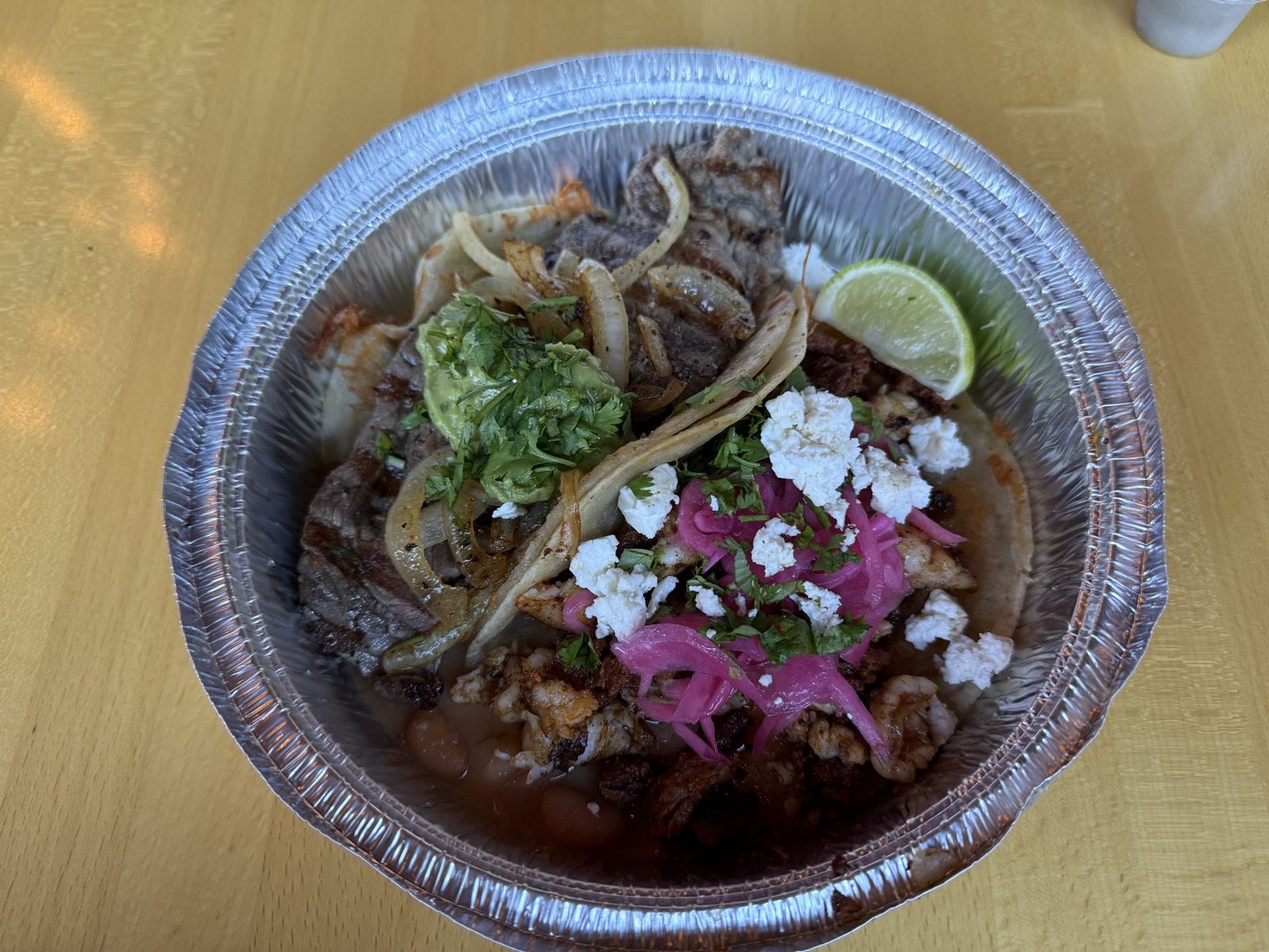 A foil takeout tray of carne asada tacos from Tacos Fita at The Heist, topped with pickled red onion, crumbled cotija cheese, guacamole and cilantro, with grilled onions, a bean side and a lime wedge