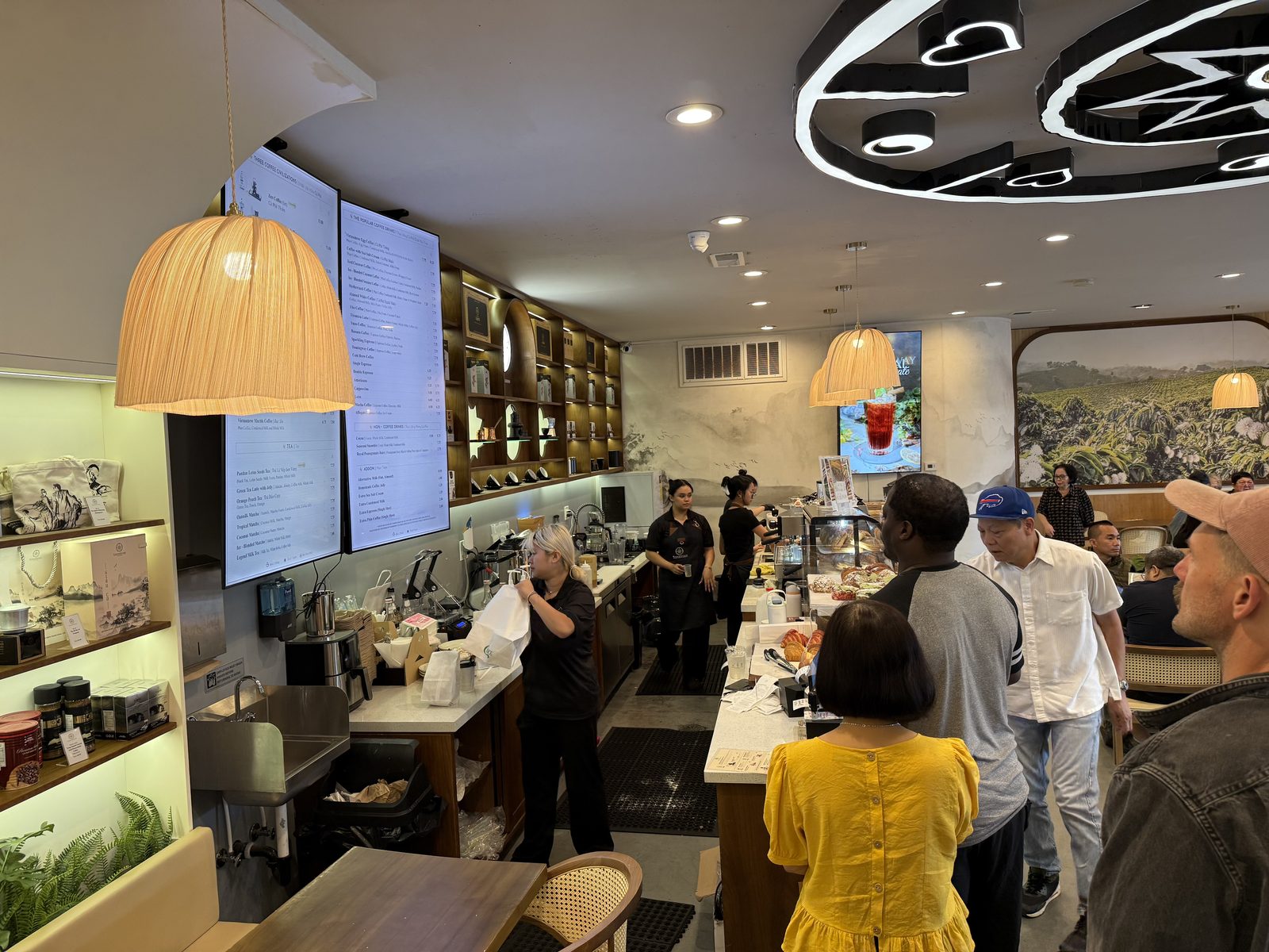 The order counter at Trung Nguyên Legend, with digital menu screens overhead, pendant lamps, a pastry case, and a line of customers waiting to order