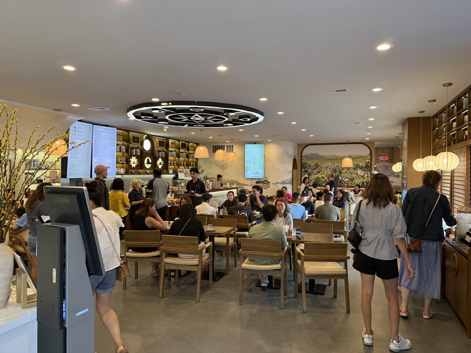 A full dining room at Trung Nguyên Legend — wood tables under a black circular ceiling medallion, digital menu screens on the back wall, a mural of a coffee farm, and a line of customers at the counter