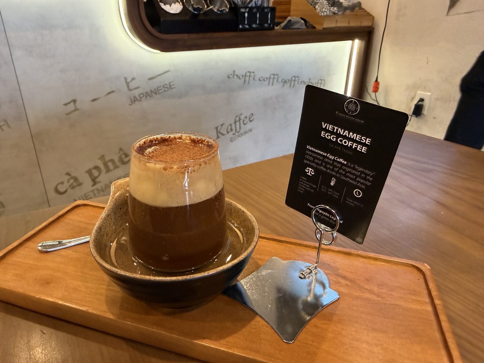 A glass of Vietnamese egg coffee — dark coffee topped with a thick pale whipped foam dusted with cocoa — sitting in a dark ceramic bowl of hot water on a wooden tray, with a small tented card reading Vietnamese Egg Coffee