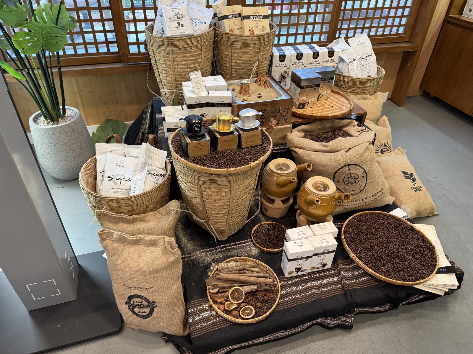 A retail display of Trung Nguyên coffee products — woven baskets of whole coffee beans, burlap sacks stamped with the compass logo, boxes of drip-filter packets, and a dish of cinnamon sticks and dried orange slices