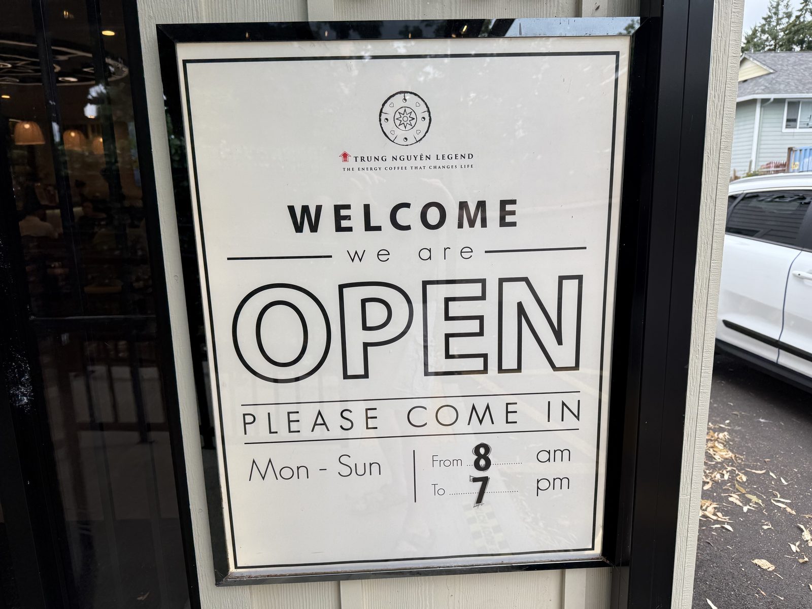A framed door sign reading Welcome, we are OPEN, please come in, Mon–Sun from 8am to 7pm, under the Trung Nguyên Legend logo and the tagline the energy coffee that changes life