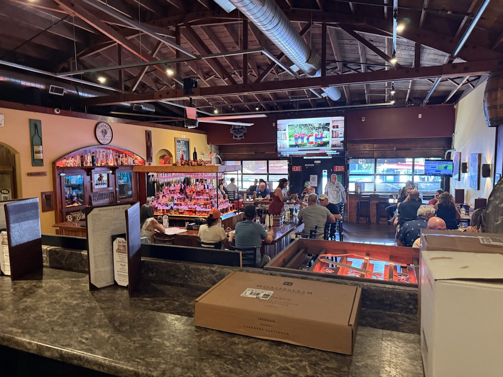 The main room of the Uptown Barrel Room under a high open-truss ceiling with exposed ductwork, a long bar with backlit liquor bottles and hanging glasses, wine-themed murals on warm yellow and terracotta walls, televisions above, and diners seated along the bar and at tables on a busy weekday evening