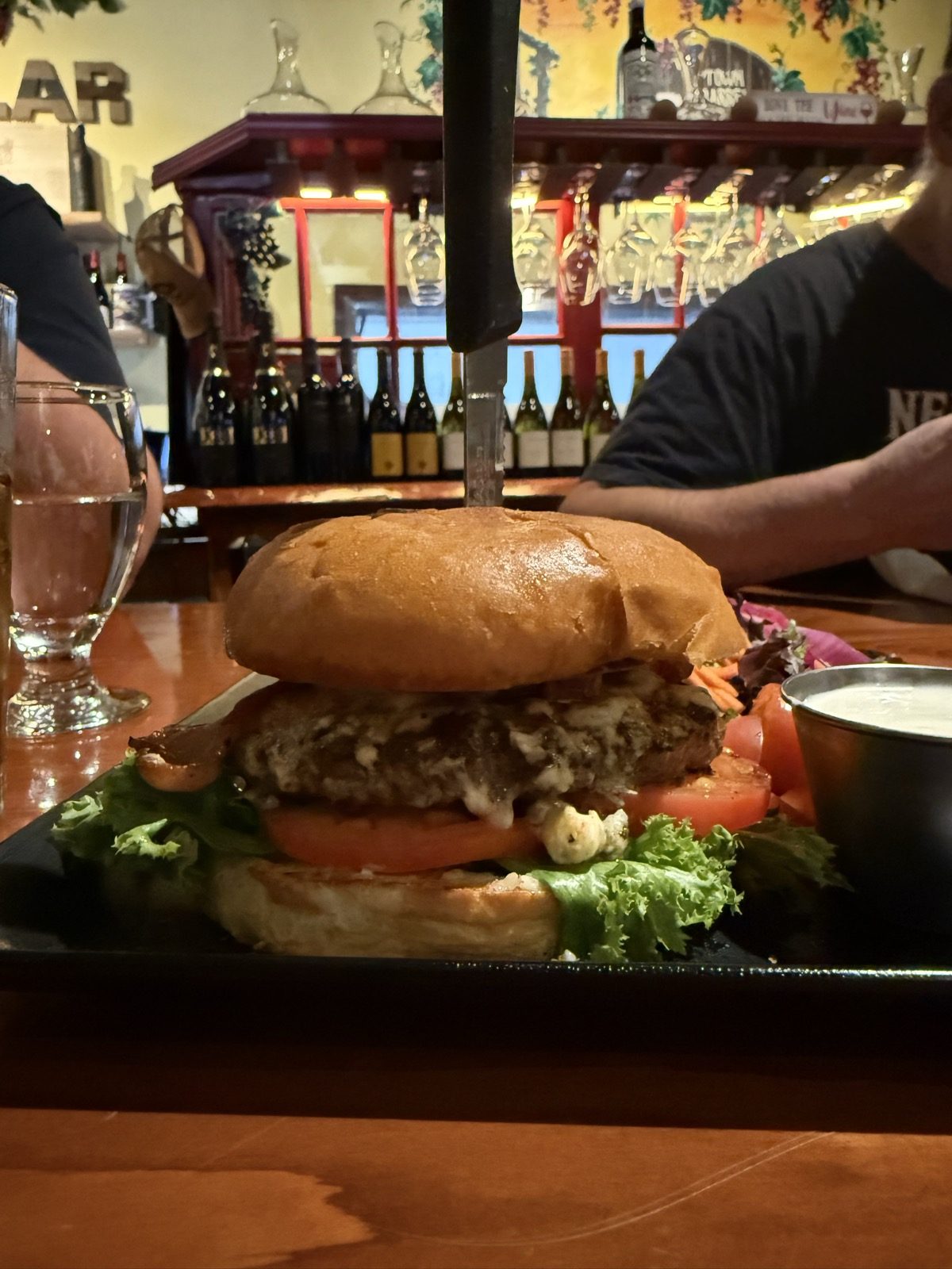 A close-up of the Black and Bleu Burger at eye level — a thick blackened Angus patty with white bleu cheese melting over the edges and crumbling onto the plate, bacon and lettuce beneath, a thick tomato slice, on a soft egg-washed bun with a black-handled knife driven down through the center, against a backdrop of the restaurant's lit wine racks and hanging glassware