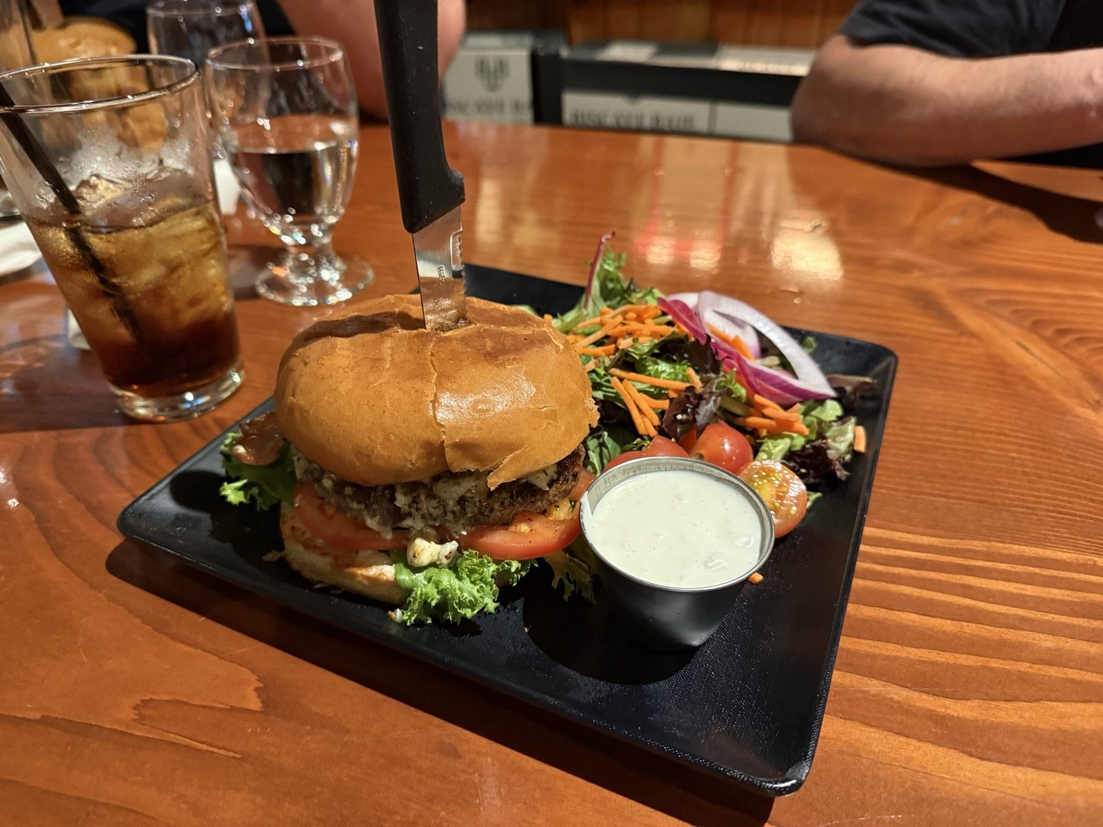The burger from above on a square black plate with a side salad of spring mix, shredded carrot, red onion and cherry tomatoes, a small metal cup of ranch dressing, and a glass of cola on the polished wood bar top