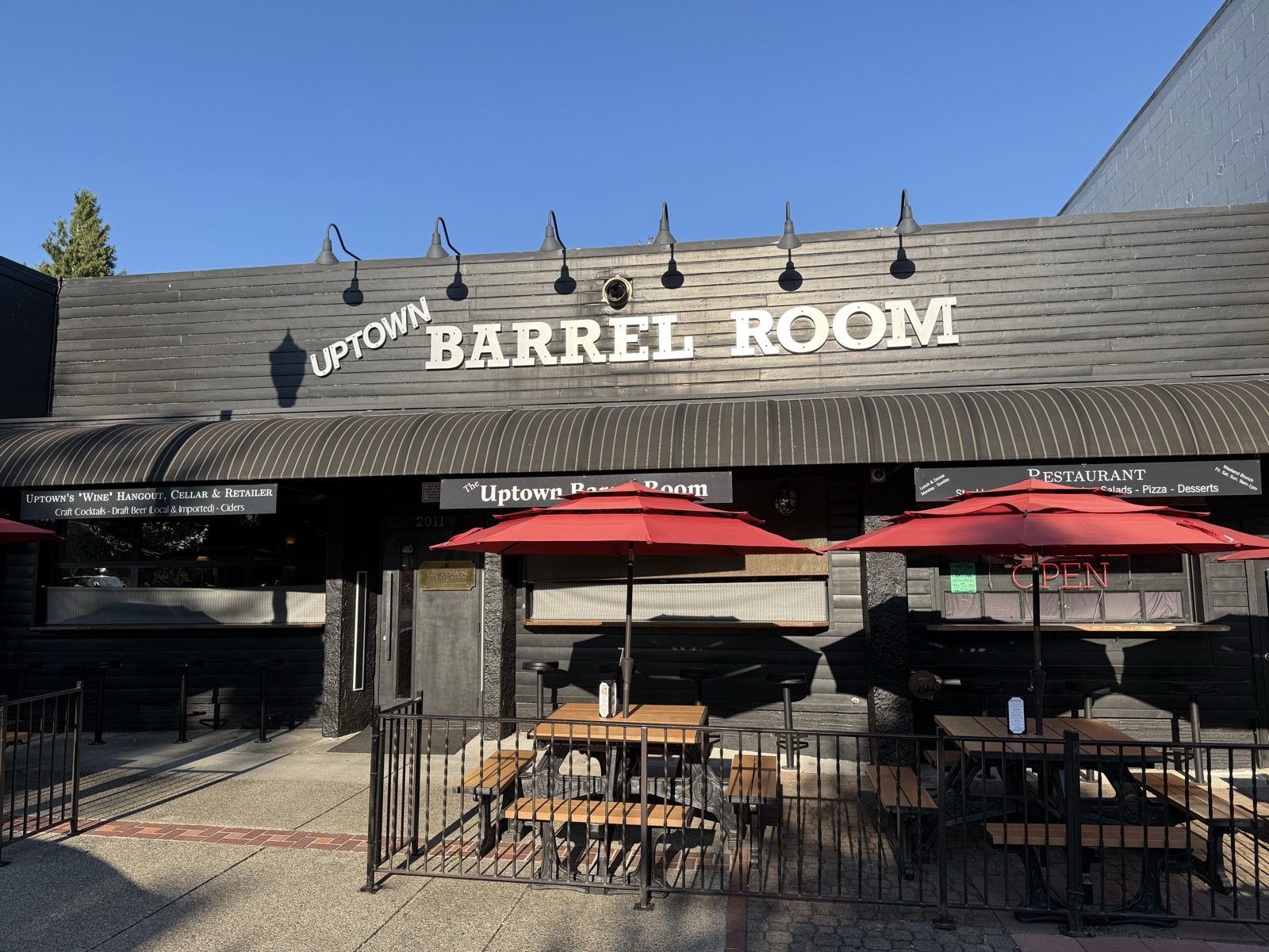 The Uptown Barrel Room on Main Street in Vancouver, Washington, photographed from the sidewalk in evening sun — a low black wood-clad building with UPTOWN BARREL ROOM in tall white letters across the front under a row of gooseneck lamps, a striped awning, a red neon OPEN sign in the window, and a fenced sidewalk patio of picnic tables under red umbrellas