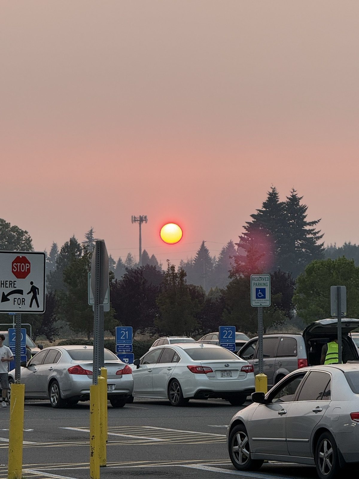 A deep red-orange sun low in a flat peach-coloured smoky sky above a line of dark fir trees, seen across a supermarket parking lot with numbered grocery pickup stalls, parked cars and a person in a hi-vis vest at an open hatchback